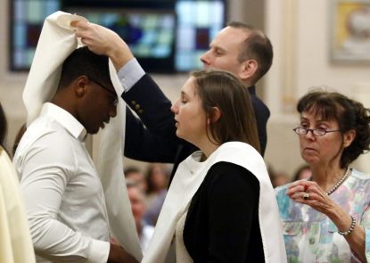 The baptismal sponsors of Christian Smith and Natalie Coughlin place baptismal garments on them as they are received into the Catholic Church. (Sarah Webb)